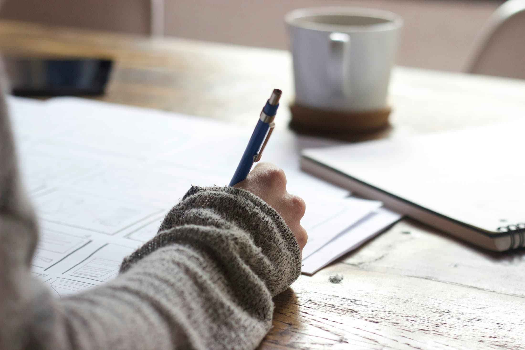 A hand writing with a pen in a notebook, a cup of coffee next to it.