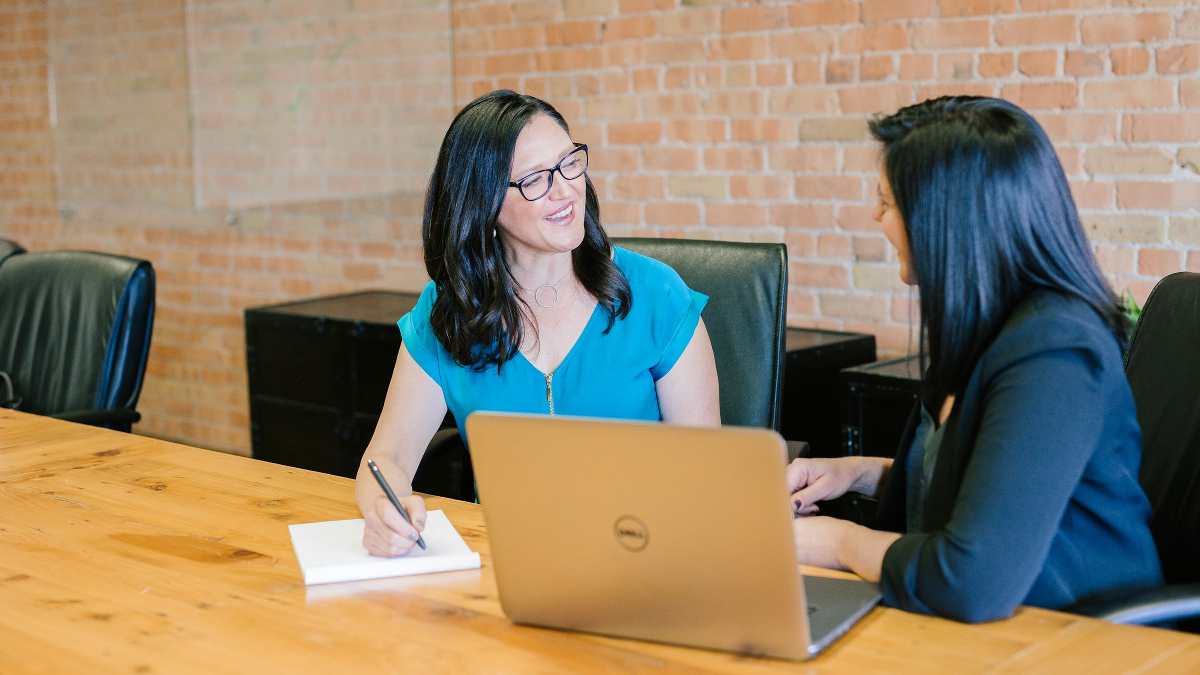 Two colleagues talking at a table, one writing in a notebook.