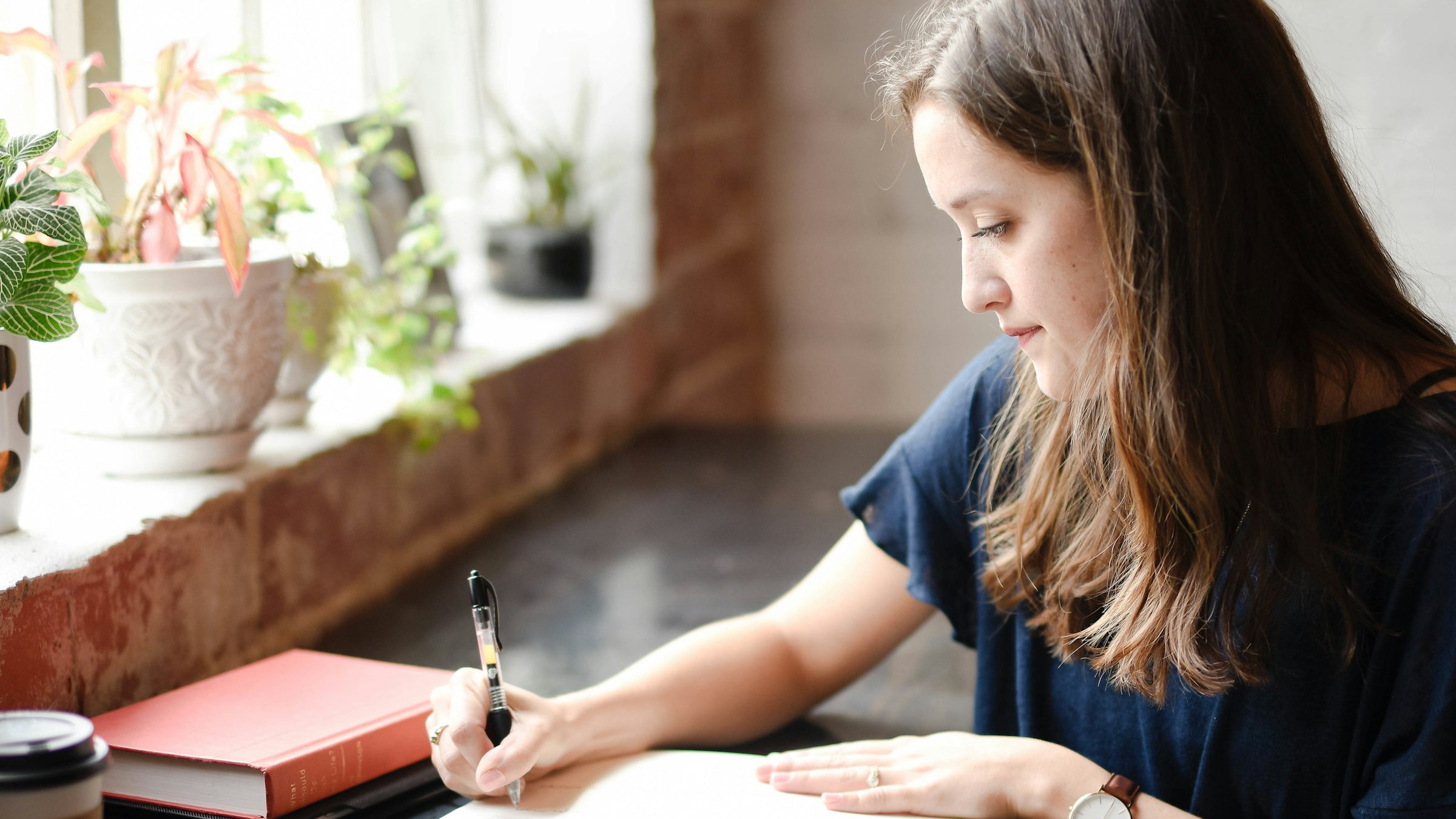 A woman writing in a notebook at a bright window table.
